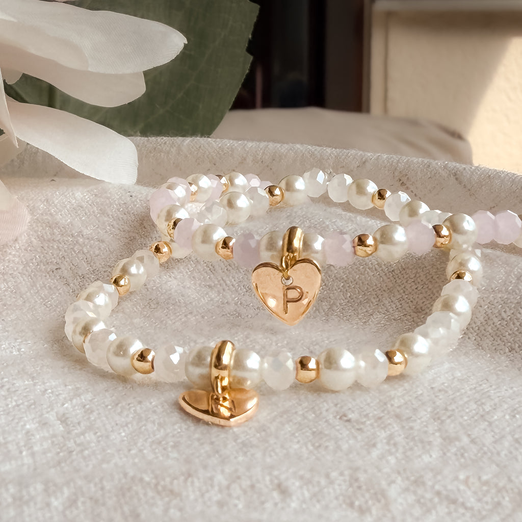Personalisiertes Perlenarmband mit Herz-Anhänger in den Farbvarianten Weiß und Rosa aus Muschelperlen, Glasperlen und vergoldeten Details.
